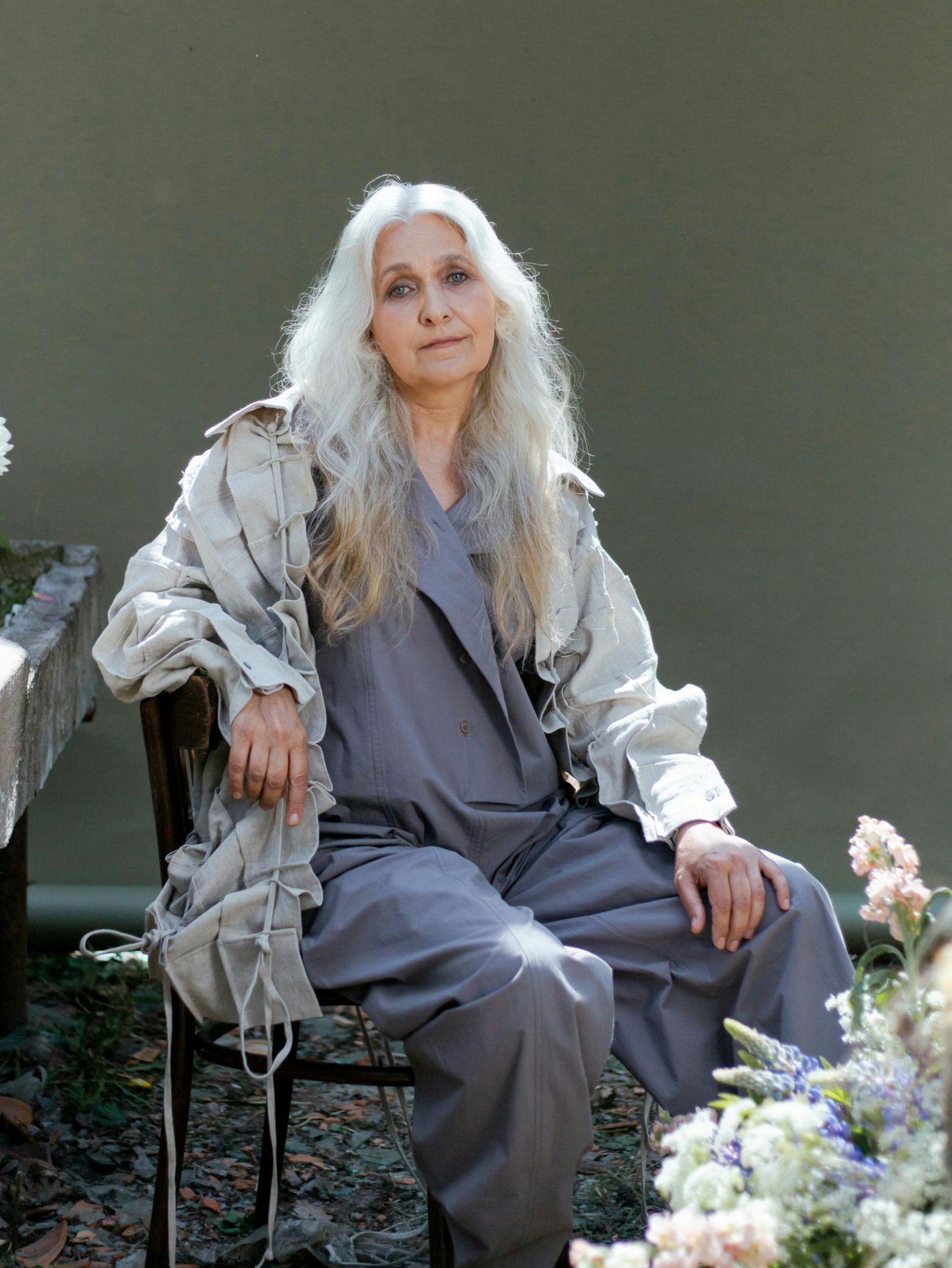 Woman with long white hair, wearing grey jumpsuit and tan jacket, seated outside, looking at the viewer.