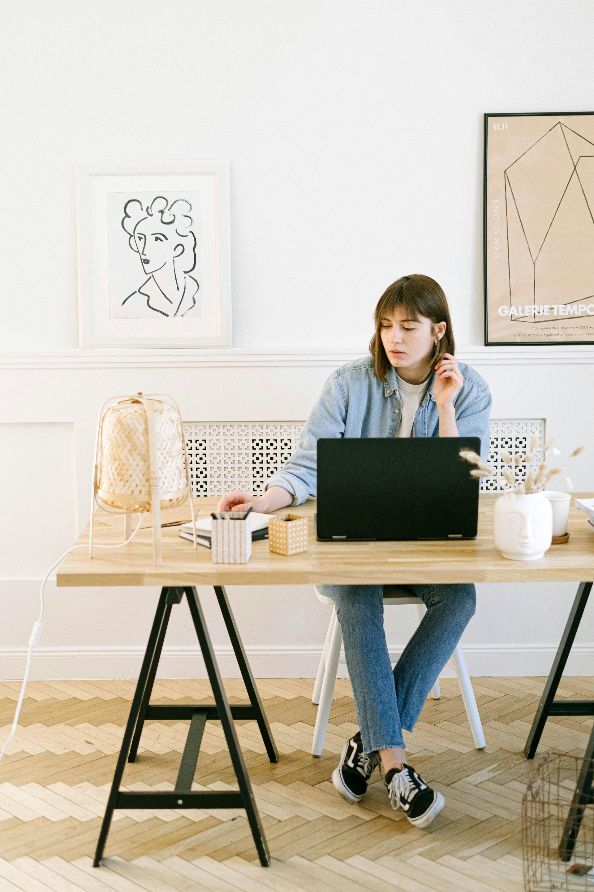 Woman in denim jacket and jeans, working on a laptop at a desk, in a bright room with art.