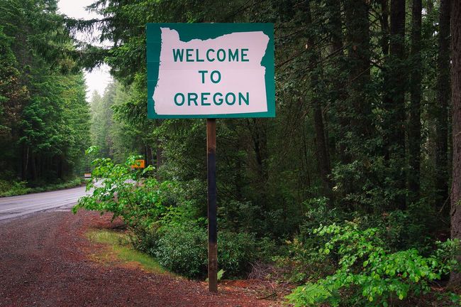 Welcome to Oregon sign with the state's outline, surrounded by trees.