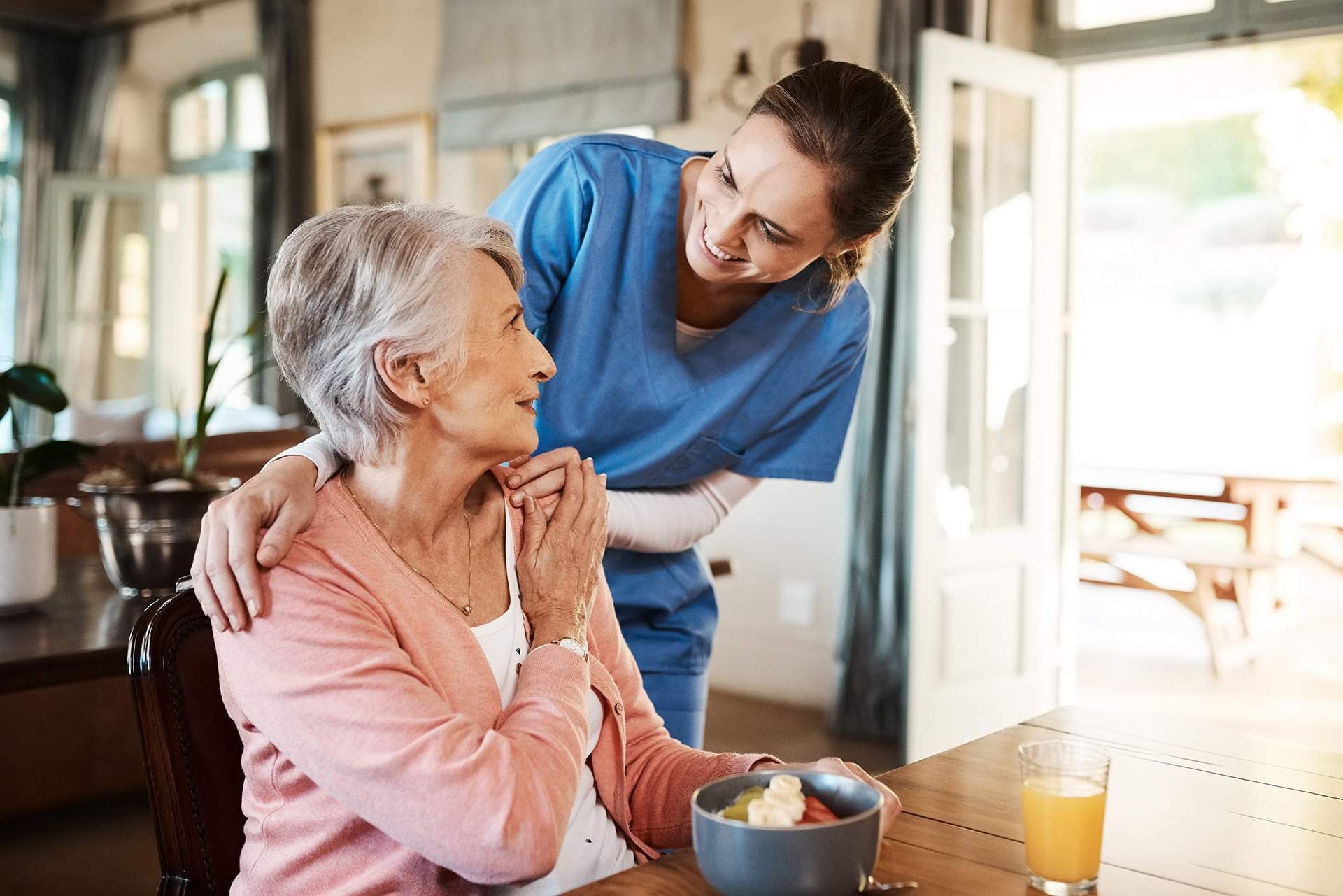 Caregiver leaning over and smiling at an older woman seated at a table in a bright home kitchen