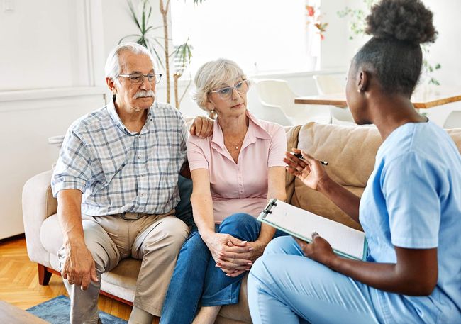 Therapist speaking with a seated couple in a bright living room, holding a notebook.