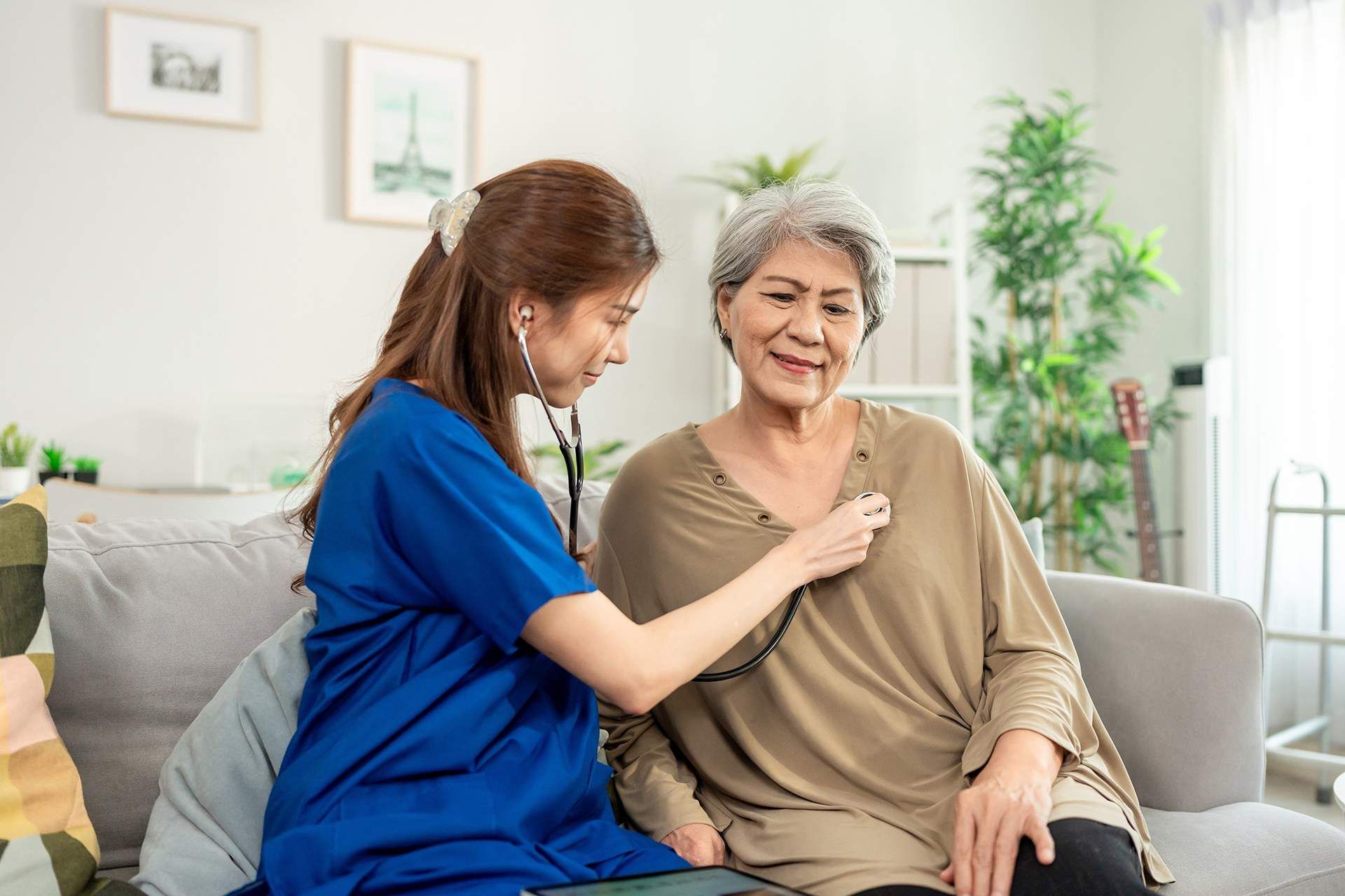 Caregiver in blue helping smiling woman with a chest stethoscope check on a sofa in a bright living room