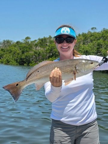 Woman smiling, holding a redfish she caught on a boat, blue water and mangrove trees in background.