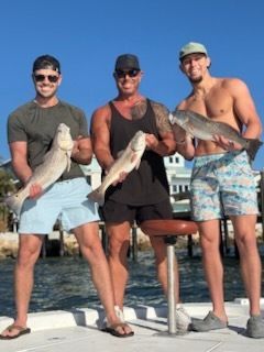 Three men holding fish on a boat; sunny day, blue water, shorts, one shirtless.