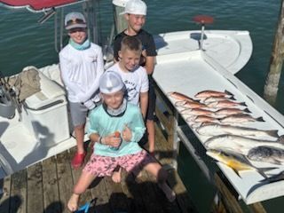 Four kids on a dock with a boat, posing near a fish cleaning station displaying several fish.