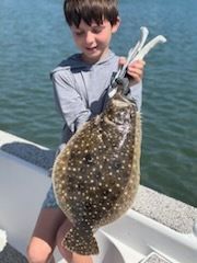 Boy holding a large, speckled flatfish with white tool in the mouth, standing near water.