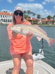 Woman holding a redfish on a boat, wearing sunglasses and a hat, in front of a waterfront building.