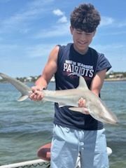 Teenager on a boat holds a small shark. He smiles, wearing a navy Patriots shirt and blue shorts, ocean in background.
