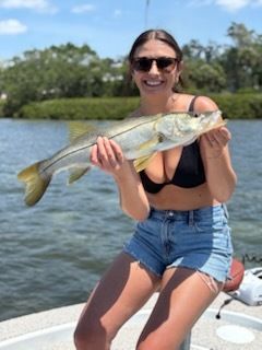 Woman in sunglasses and shorts smiles, holding a silver fish on a boat.
