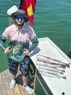 Man on dock with caught fish, wearing hat, sunglasses, fishing shirt, and shorts; ocean backdrop.