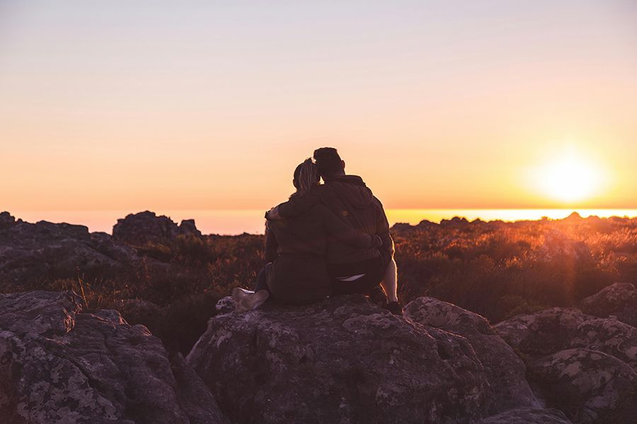 A couple is sitting on top of a rock watching the sun set over the ocean.