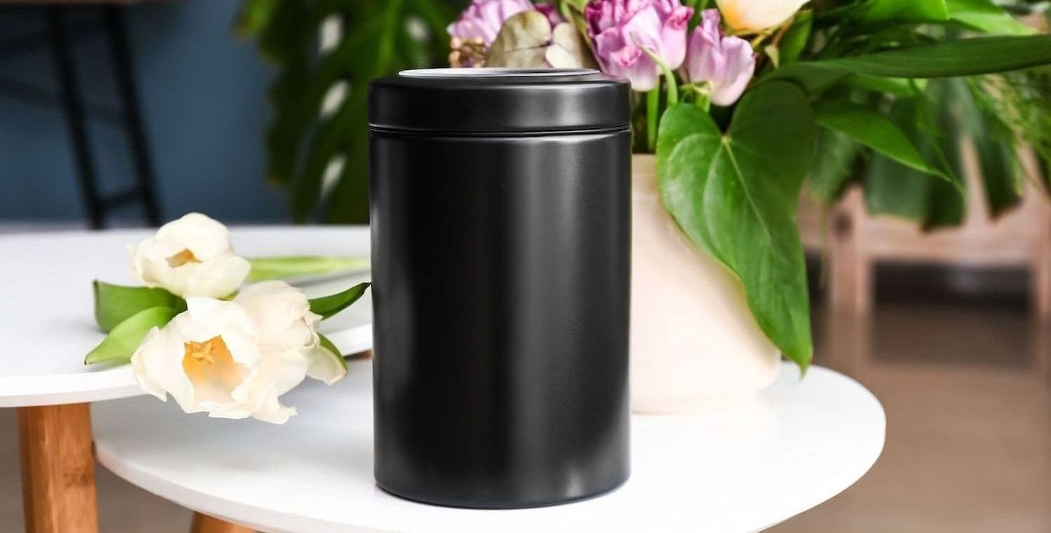 A black canister is sitting on a white table next to a vase of flowers.