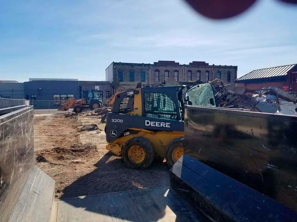 Yellow John Deere skid steer at a construction site beside rubble and a damaged building under a clear sky