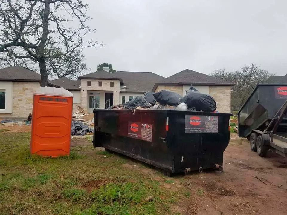 Dumpsters and an orange portable toilet on a muddy lot beside houses and trees.
