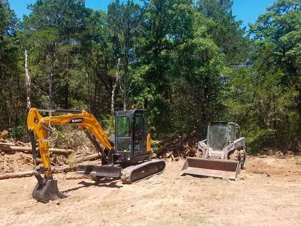 Yellow excavator and dump truck parked on a dirt clearing beside a wooded area.