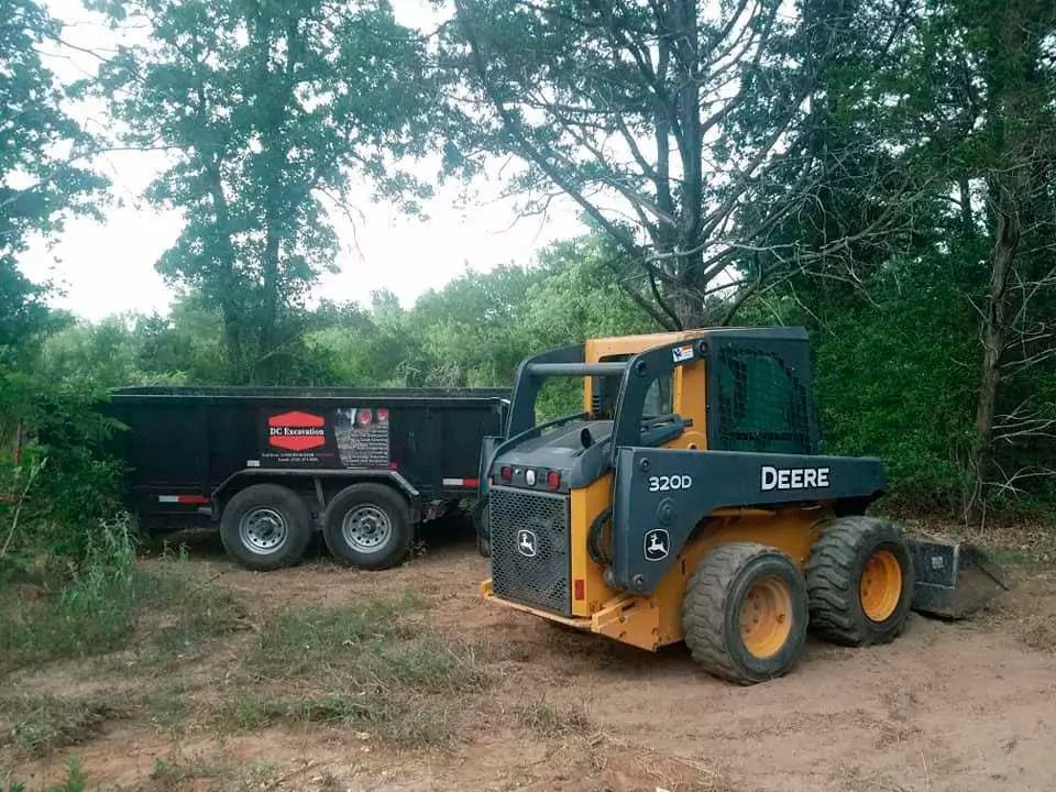 Yellow John Deere skid steer beside a black dump trailer in a wooded clearing