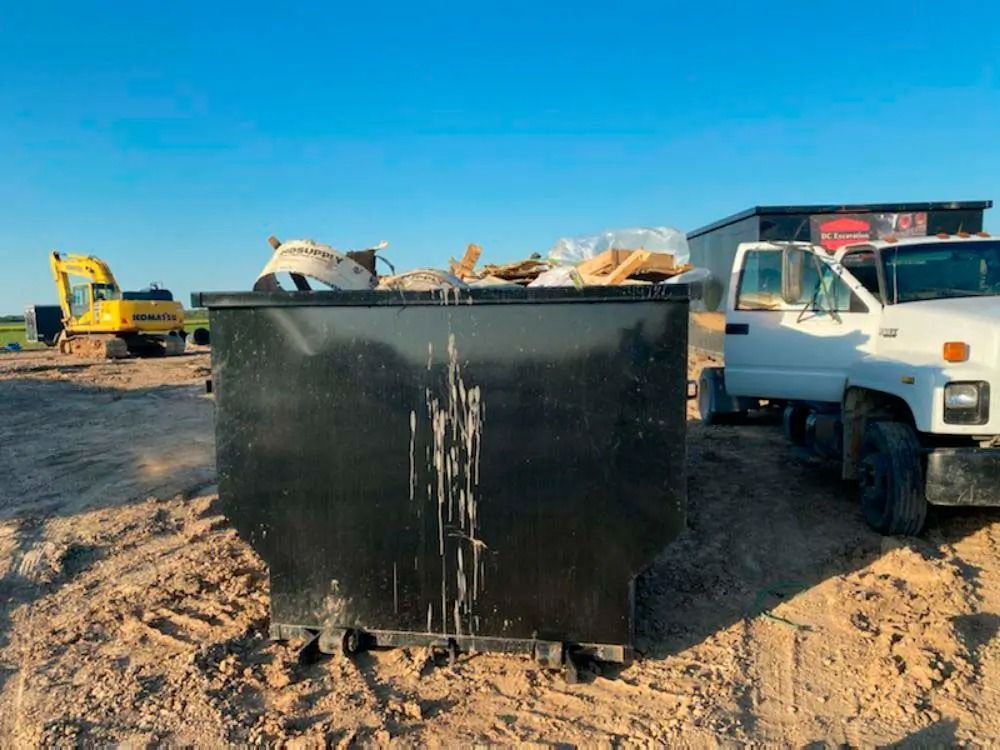Black dumpster overflowing with debris beside a white truck at a sandy construction site