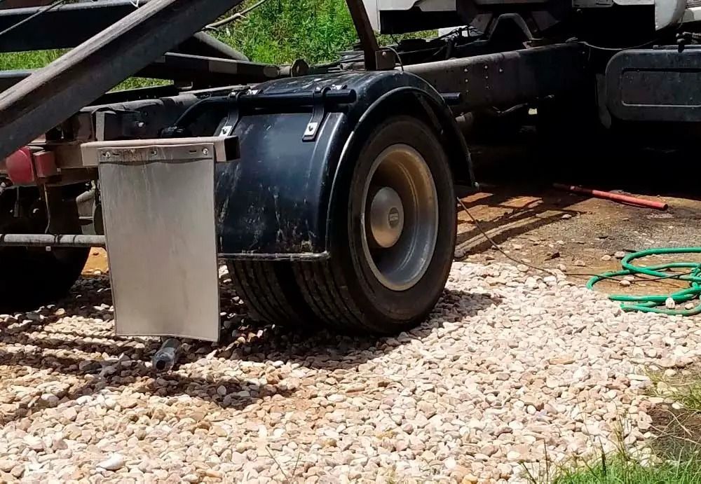 Black trailer wheel and fender on gravel driveway, with a metal panel hanging below and a green hose nearby