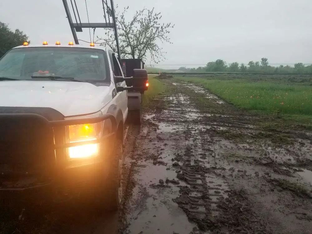 White pickup truck stuck in muddy roadside field under overcast sky, hazard lights on