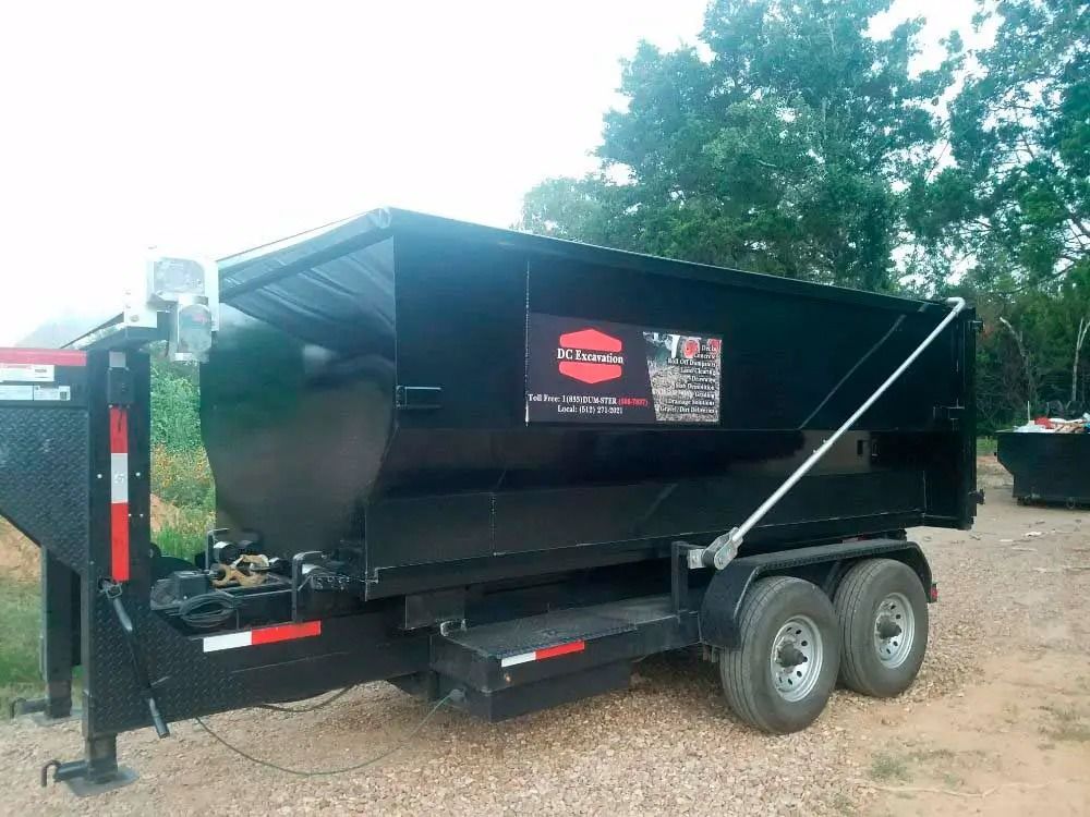 Black dual-axle dump trailer parked on dirt, with hydraulic lift and logo on the side.