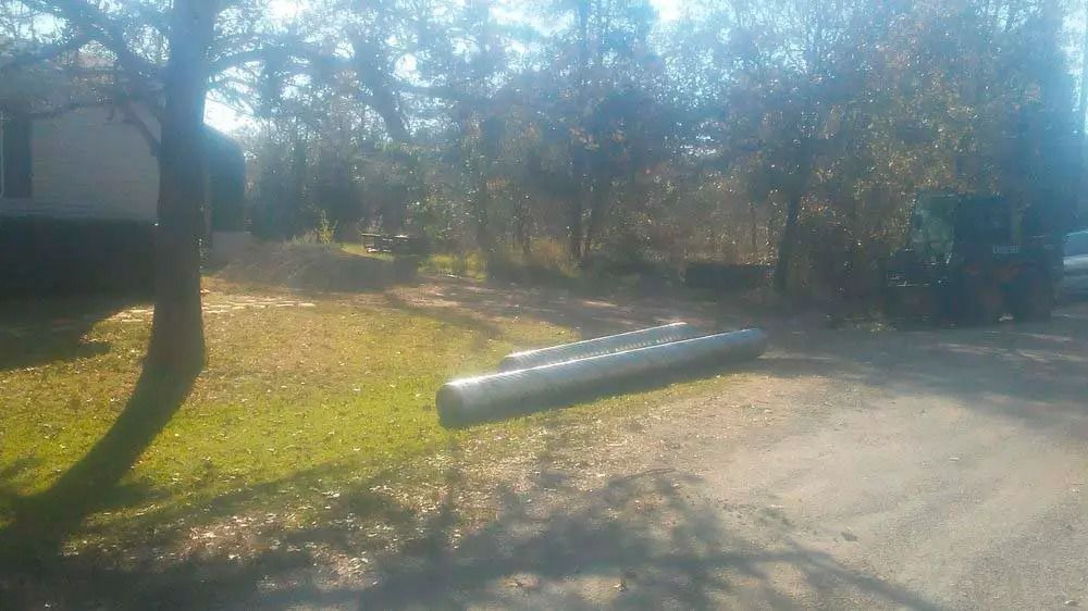 A long metal pipe lies on a grassy roadside beside trees and a dirt path.