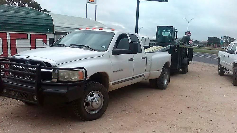 White pickup truck towing heavy equipment in a dirt lot, with another white truck nearby.