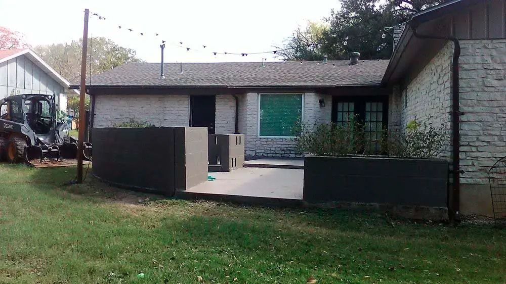 Backyard patio with stone house, covered seating area, and grassy lawn under an overcast sky