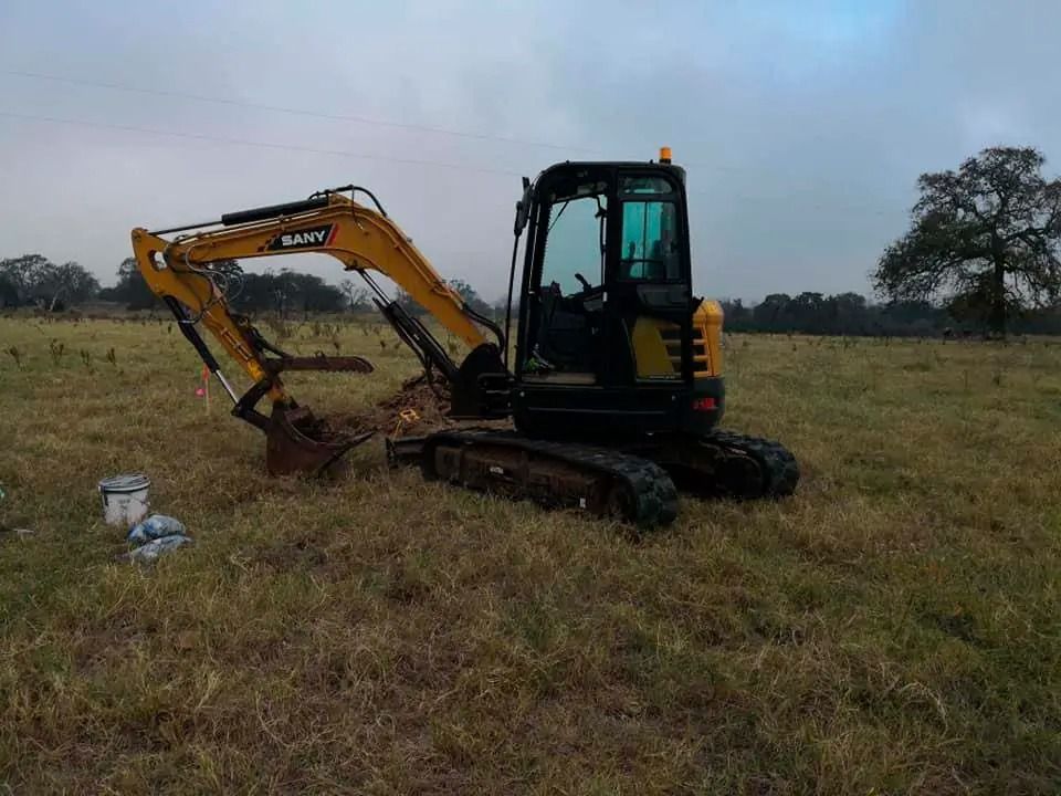 Yellow excavator in a grassy field, digging near a small concrete block under a cloudy sky