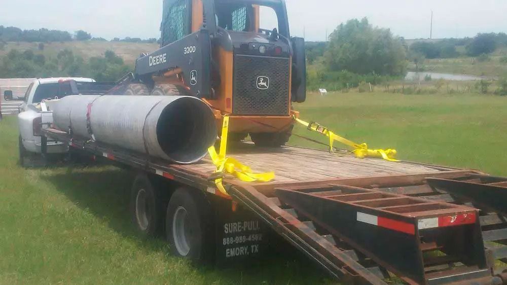 Excavator loading a large metal pipe onto a flatbed trailer in a grassy field.