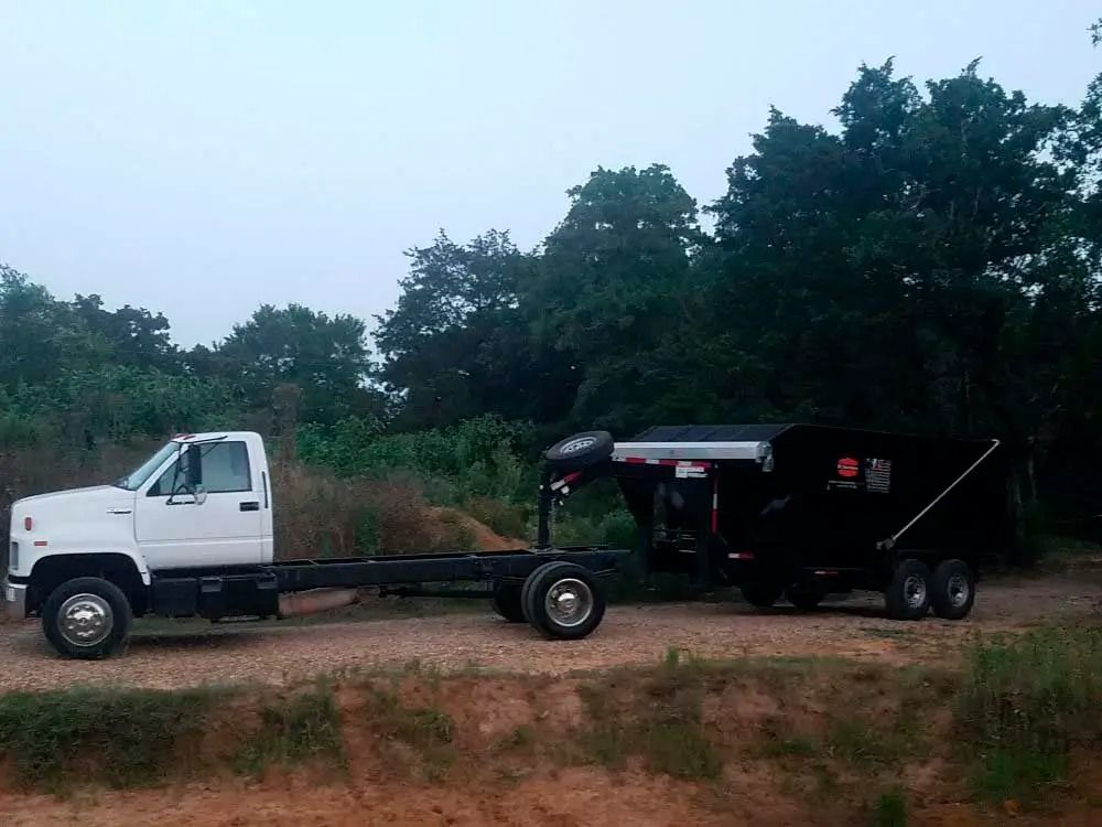 White truck towing a black dump trailer on a dirt road with trees in the background