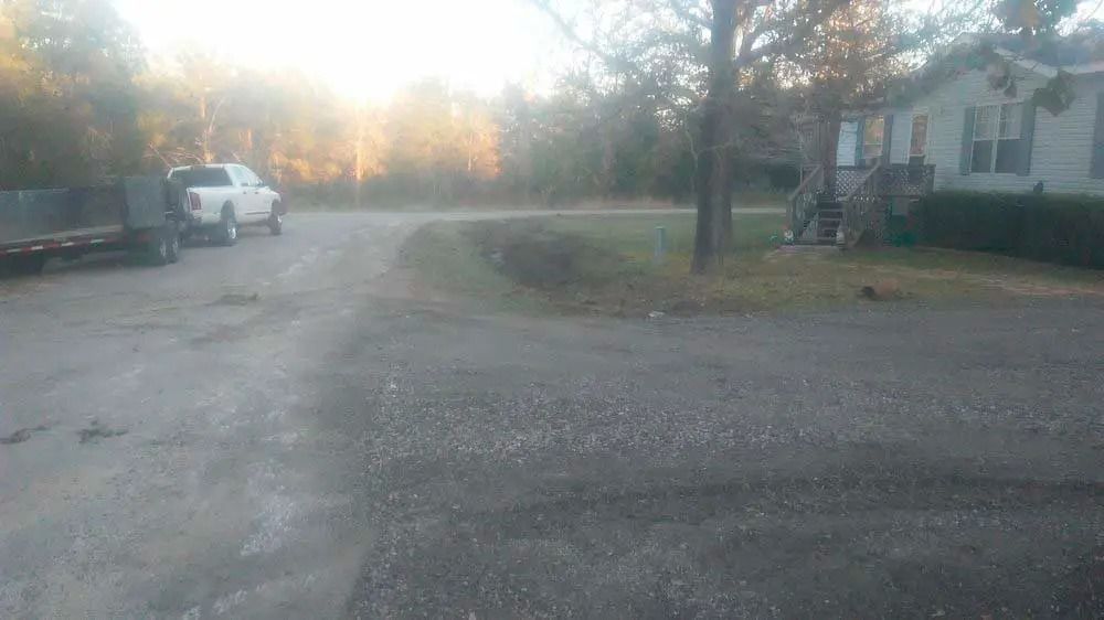 Gravel driveway at dusk with parked vehicles, trees, and a house on the right