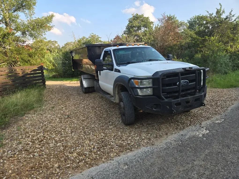 White dump truck on a gravel driveway beside trees and a wooden fence.