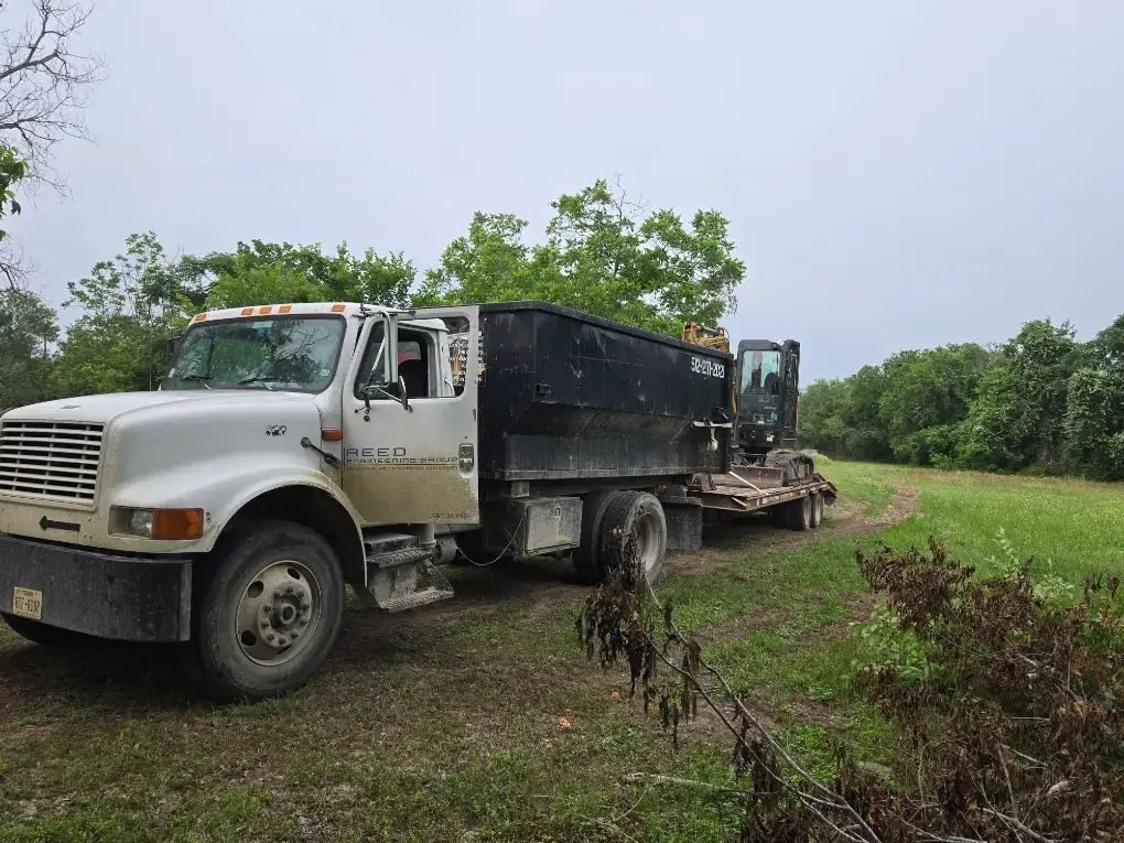 White dump truck and flatbed trailer on a dirt road beside a grassy field and trees