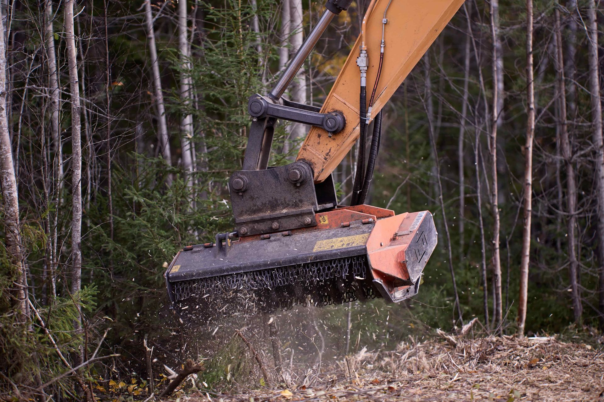 Excavator clearing brush in a wooded area, with debris flying from the attachment.