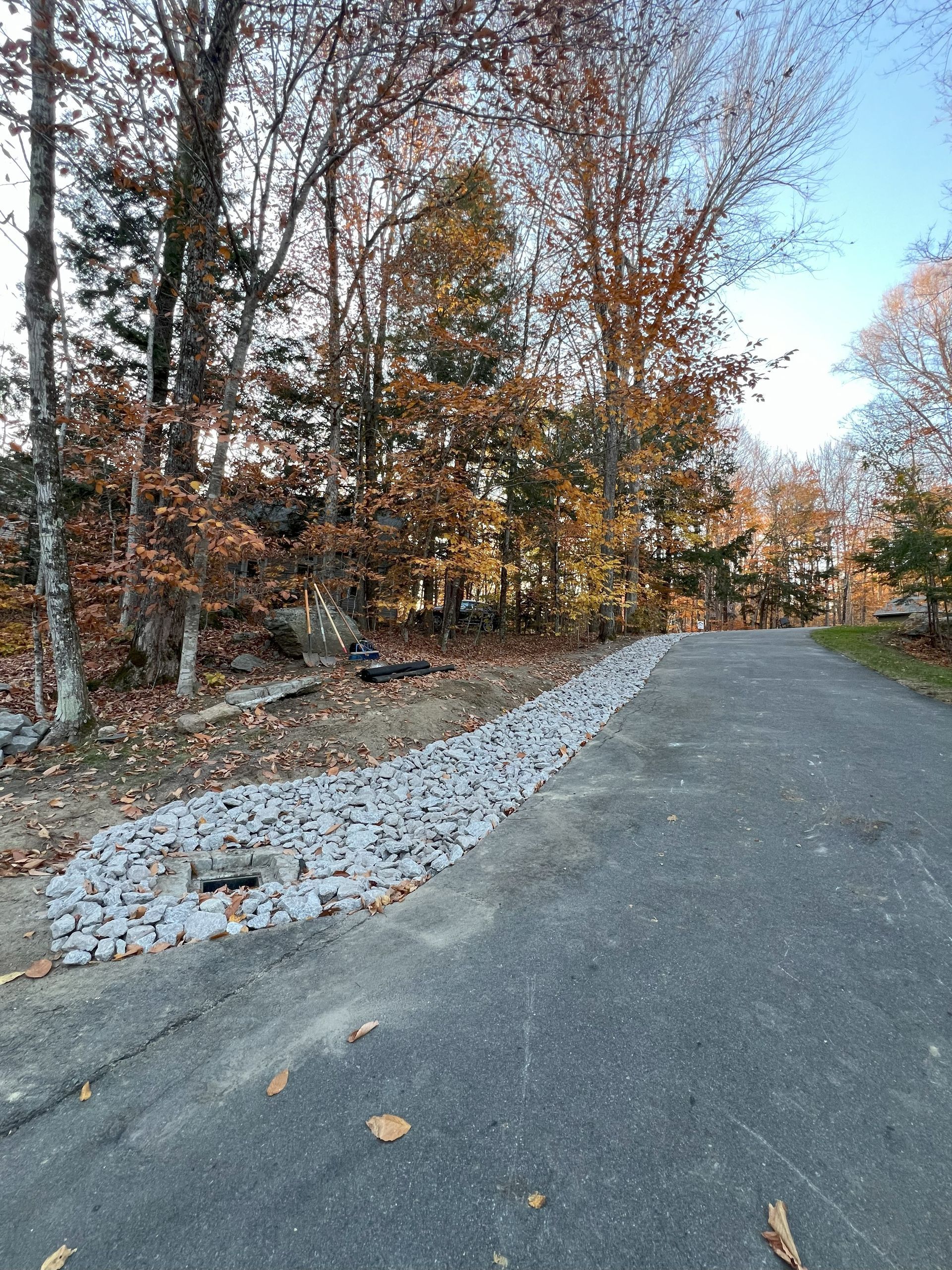 Asphalt driveway lined with gray rocks beside a wooded area with autumn foliage.