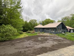 A one-story house with dark blue siding and a brown roof is surrounded by trees and a freshly tilled yard.