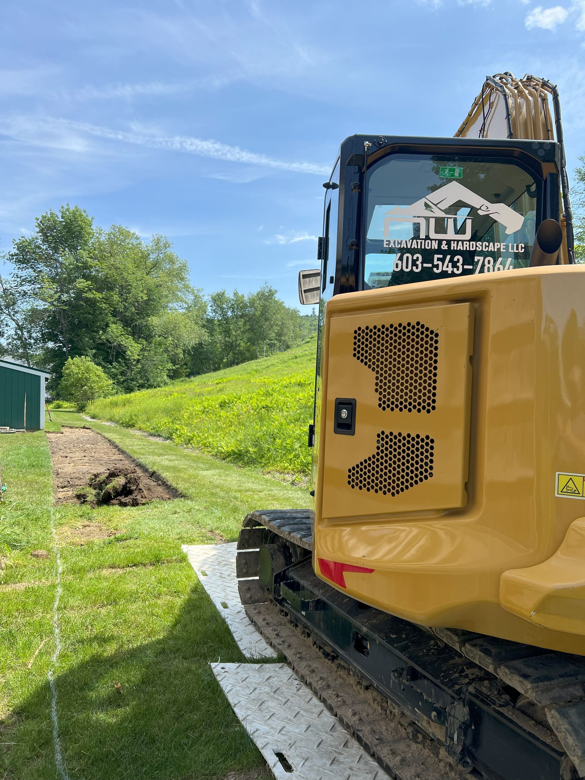 Yellow excavator digging trench in grassy area, with a tree line background under a blue sky.