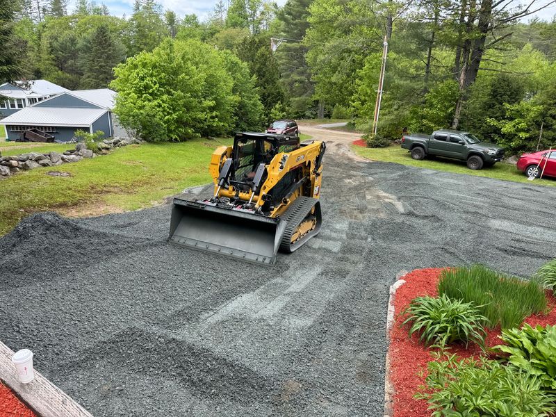 Yellow skid steer on gravel driveway, grading the surface, next to houses and cars.