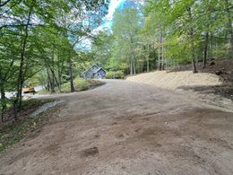 Gravel driveway leading uphill towards a small cabin in the woods. Trees surround the clearing.