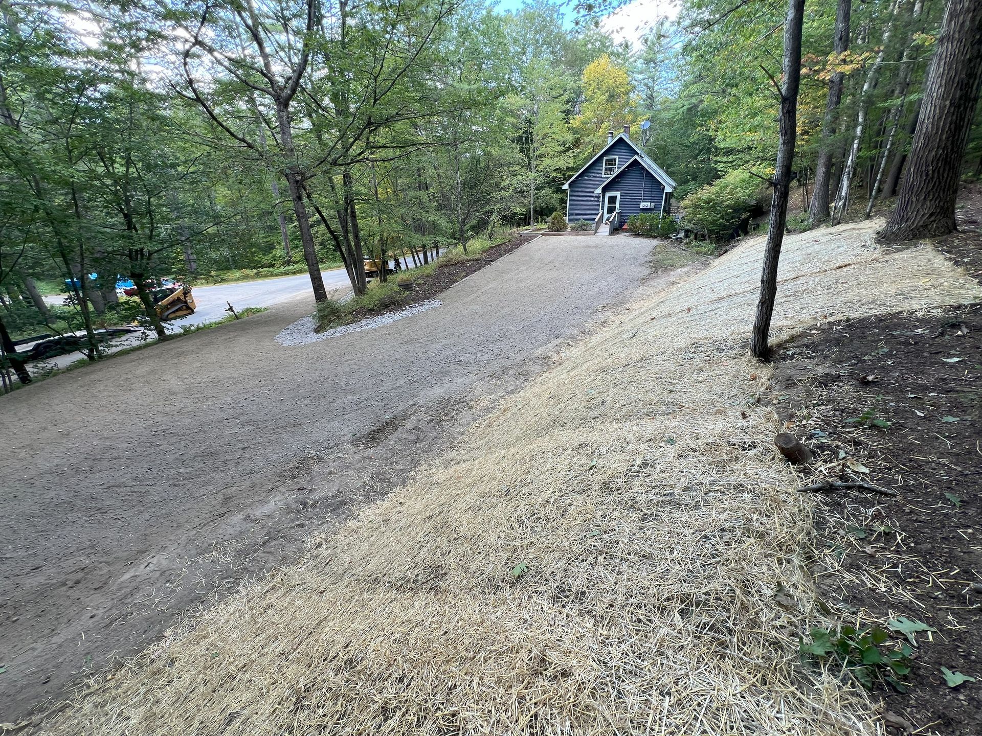 Gravel driveway leading to a small blue house on a hillside, mulched areas on either side.