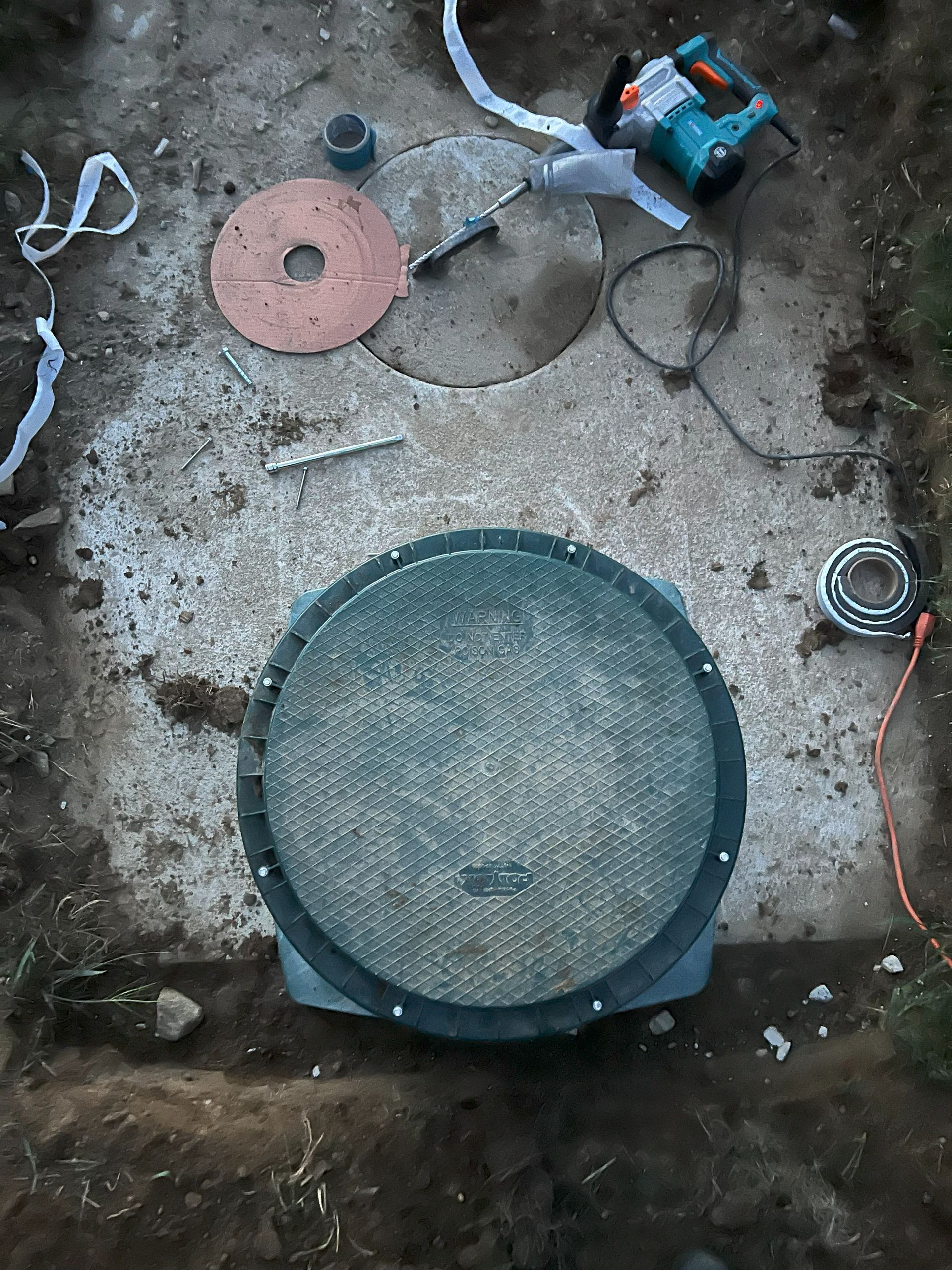 A ground-level shot of an open sewer cover in dirt, tools, and debris nearby.