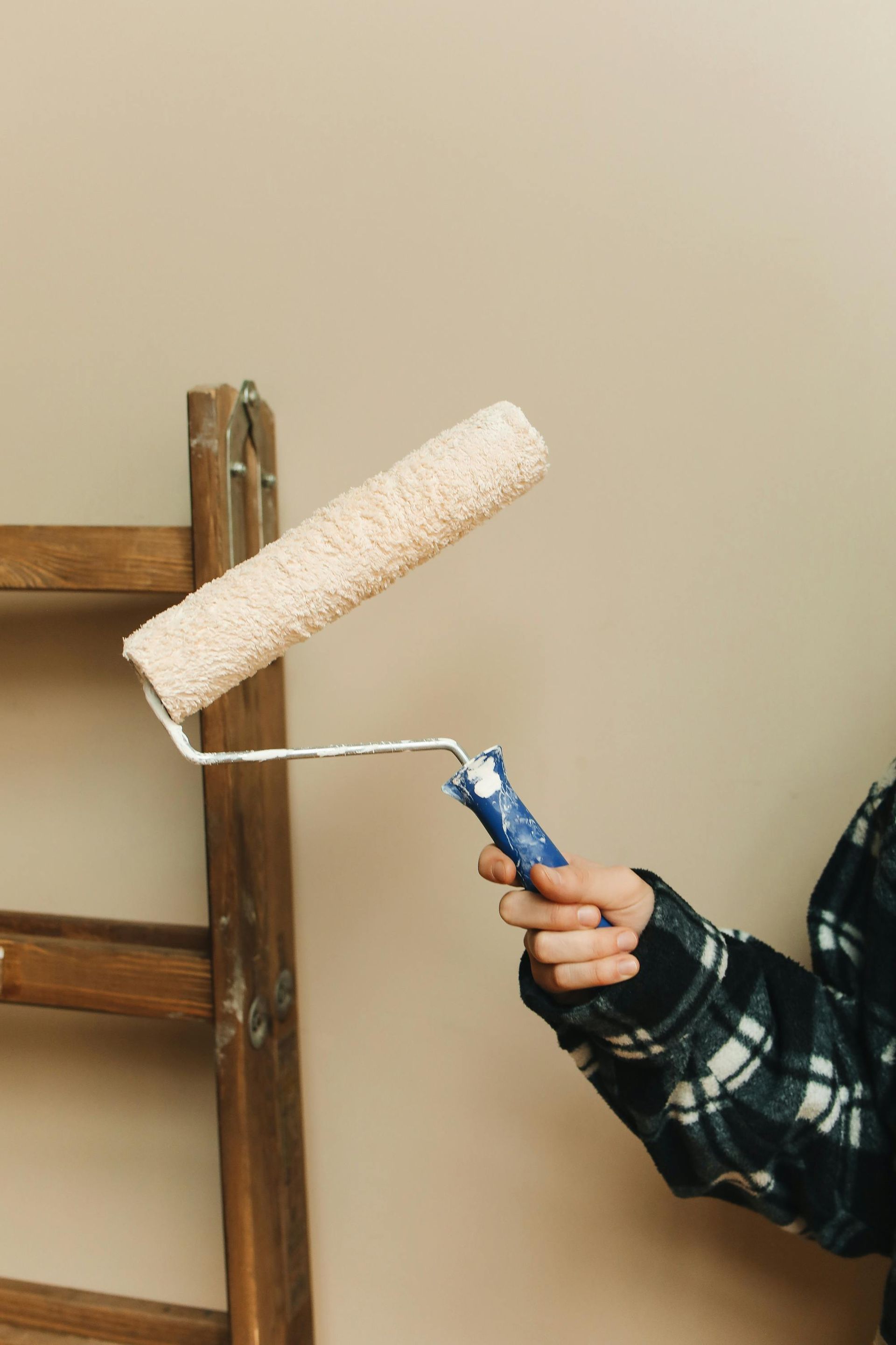 Person in plaid shirt holding paint roller near wooden ladder on tan wall.