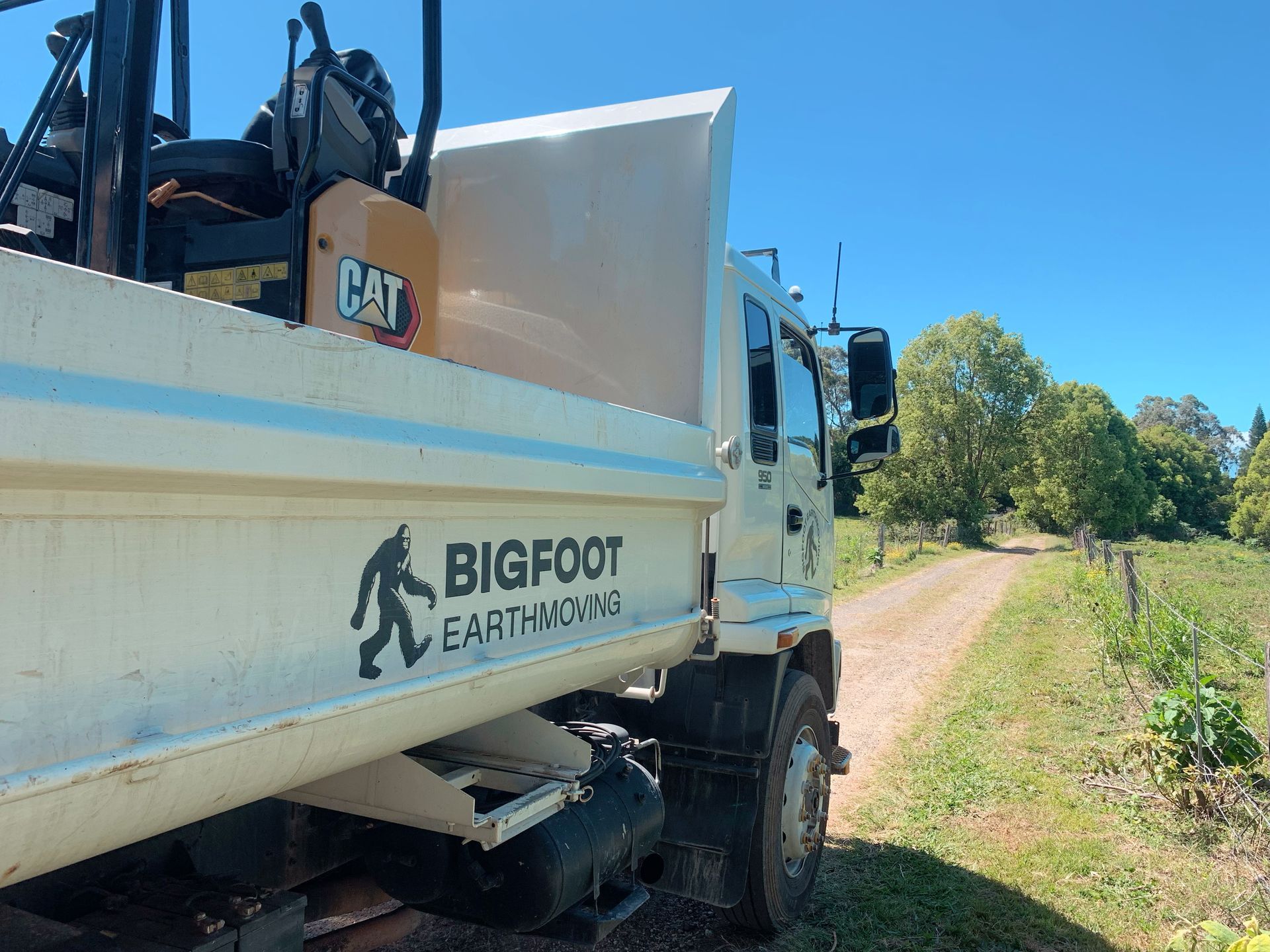 White Bigfoot Earth Moving truck on dirt road next to green trees.