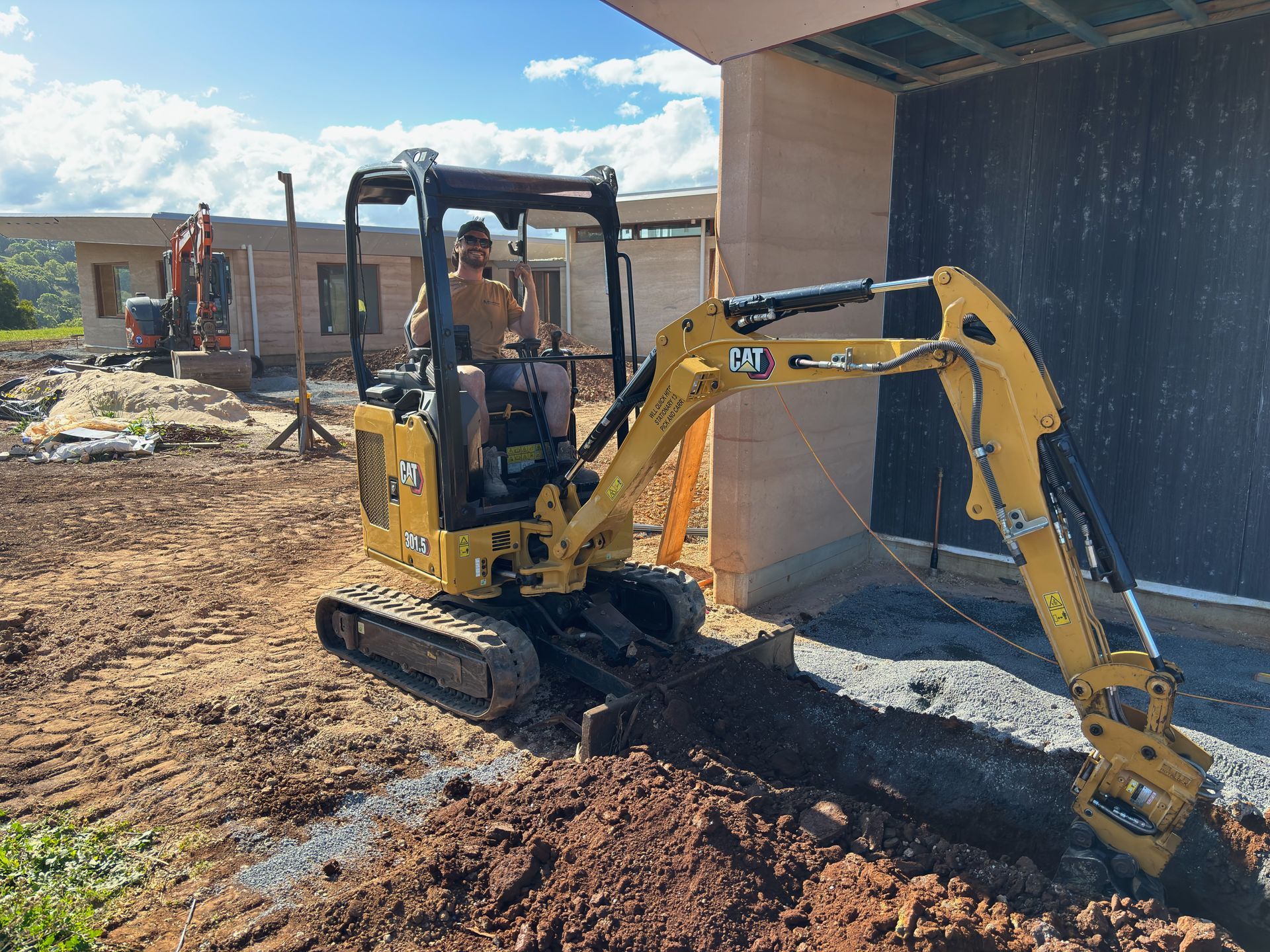Yellow excavator digging near a building, operated by a person. Outdoors, construction site.