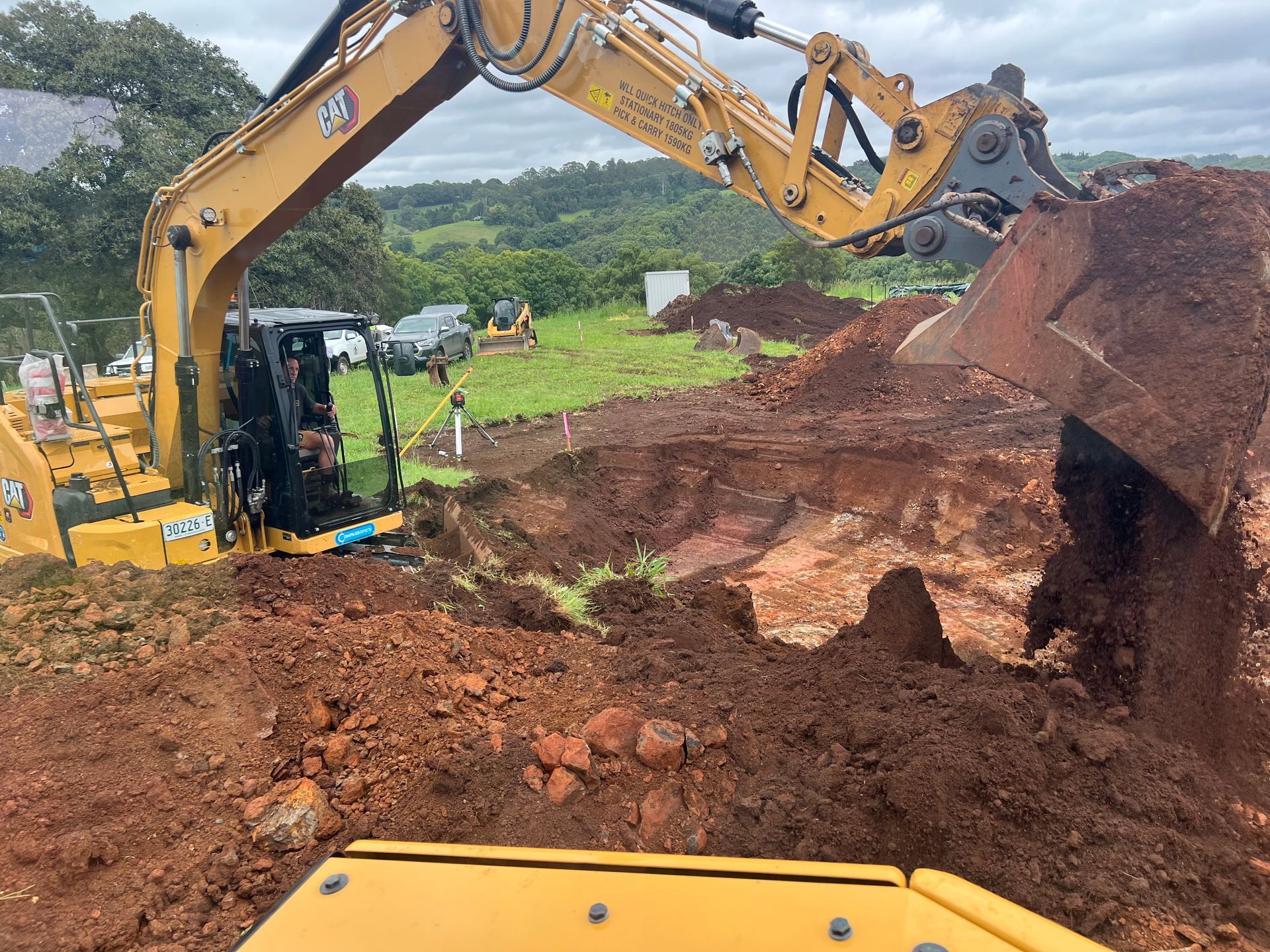 Yellow excavator digging into reddish-brown earth on a hillside, other construction equipment in the background.