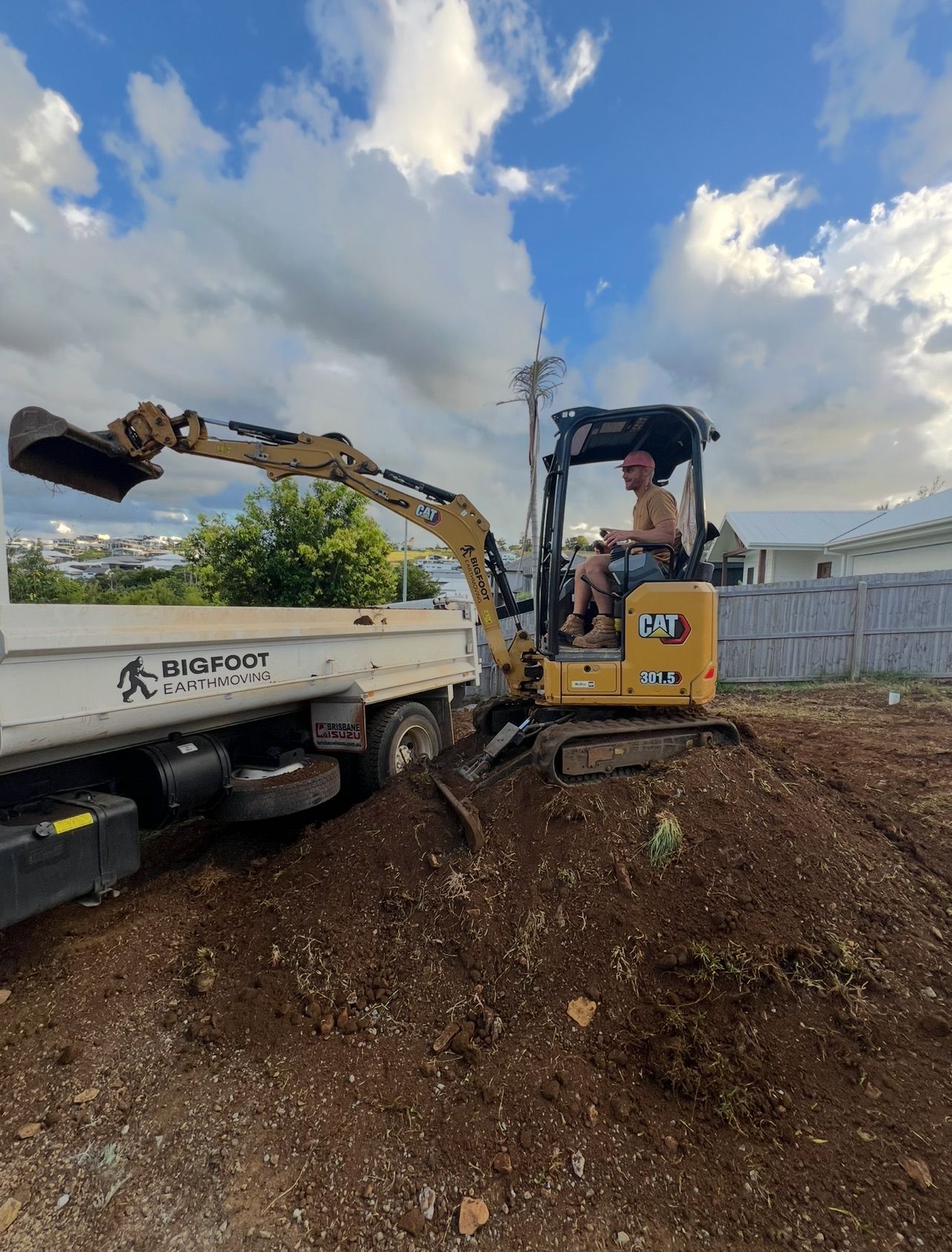 Man operating an excavator, loading dirt into a white truck. Sunny day, blue sky.