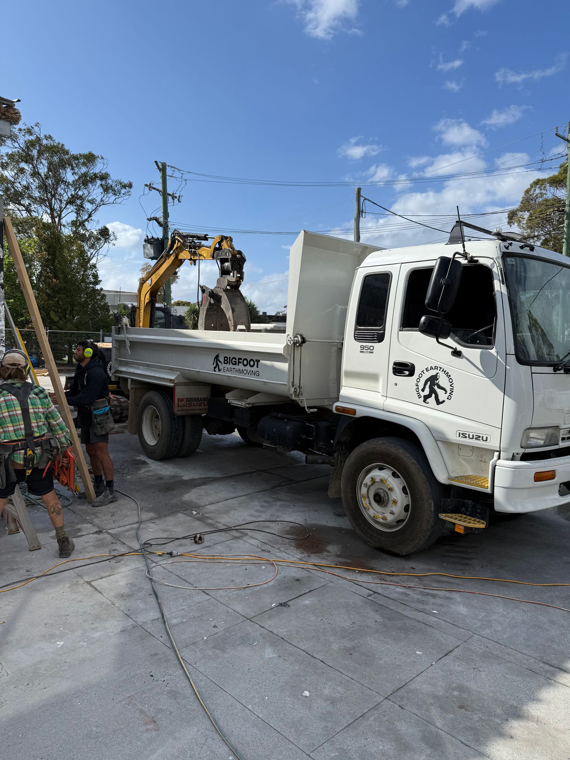 White truck with a worker operating a yellow excavator. Construction site under a blue sky.