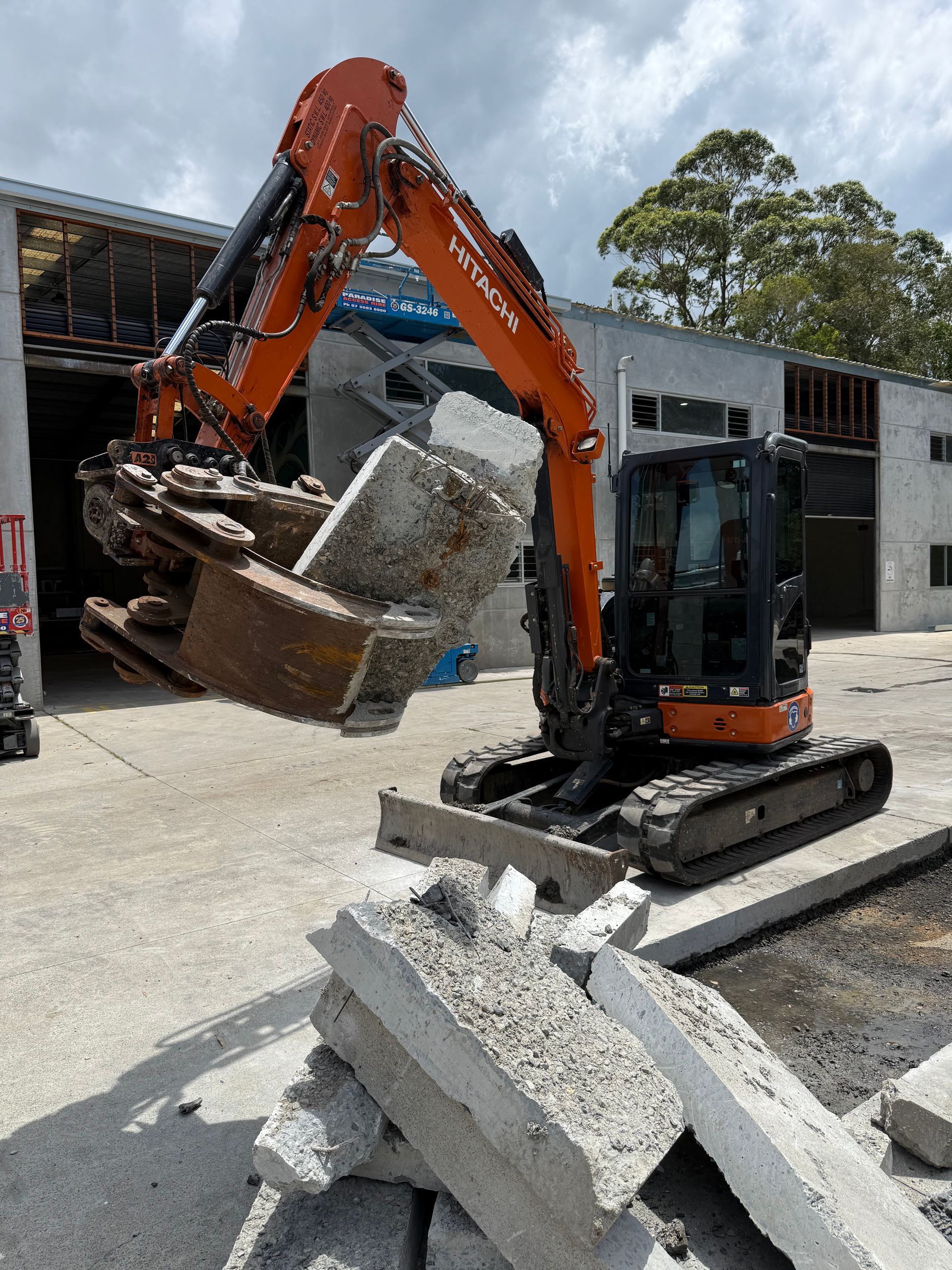 Orange excavator lifting concrete debris in front of a building with broken windows.