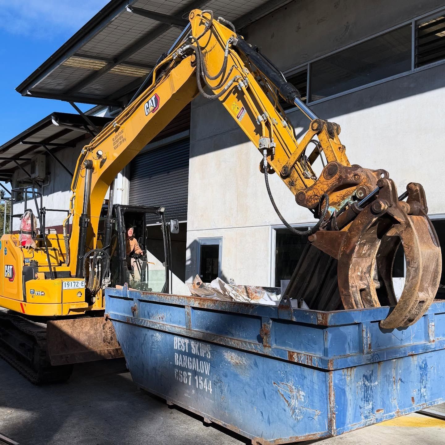 Yellow Excavator Loading Debris Into a Blue Dumpster — Bigfoot Earthmoving In Byron Bay, NSW