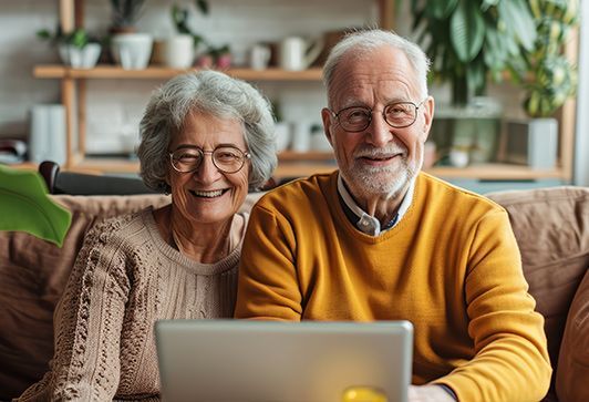 An elderly couple is sitting on a couch using a laptop computer.